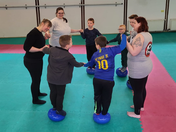 Kinder balancieren im Kreis stehend auf Bällen, unterstützt von Erwachsenen