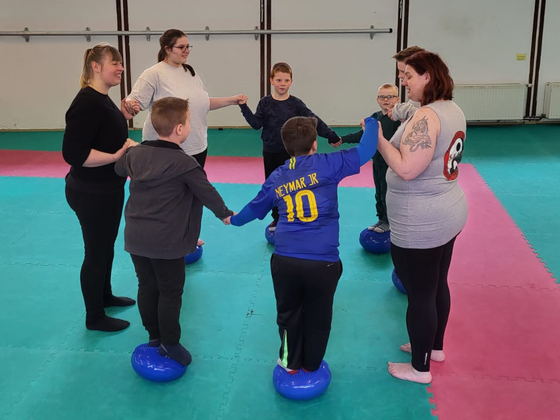 Kinder balancieren im Kreis stehend auf Bällen, unterstützt von Erwachsenen